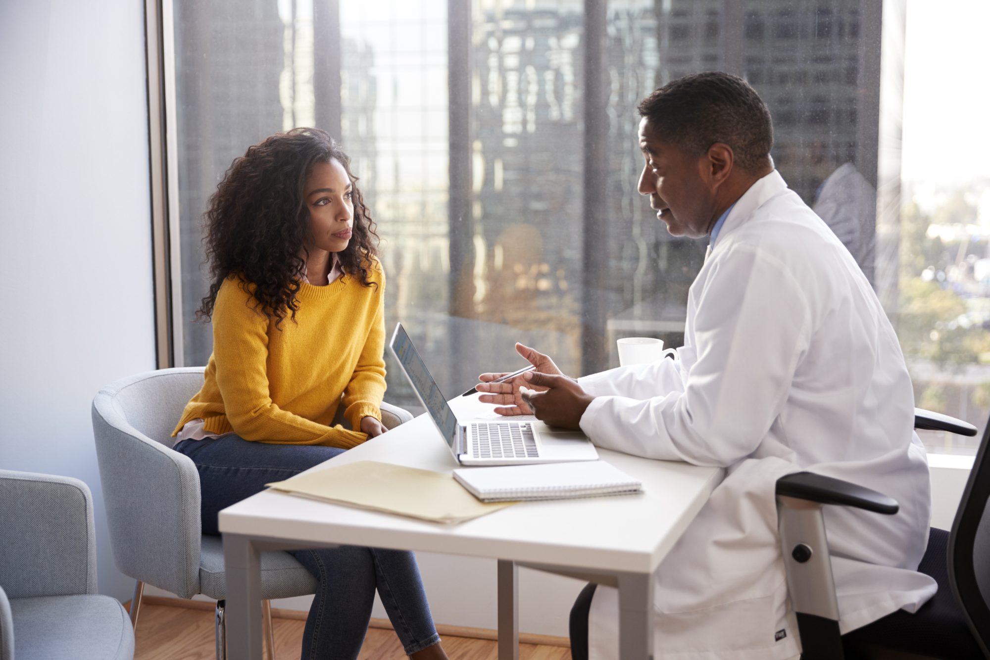 Serious Woman Having Consultation With Male NHS Doctor In Hospital Office