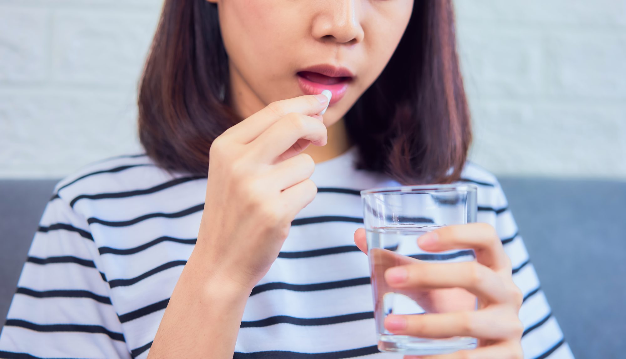 Young Asian woman in stripy tshirt taking vitamin pill with glass of water 