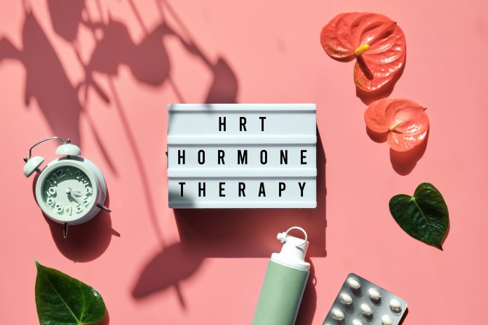 Text reads: HRT Replacement Therapy on light box. Menopause, hormone therapy concept. Pink background with alarm clock