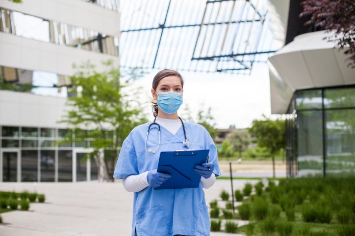 NHS worker wearing personal protective equipment holding folder standing in front of hospital