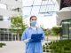 NHS worker wearing personal protective equipment holding folder standing in front of hospital