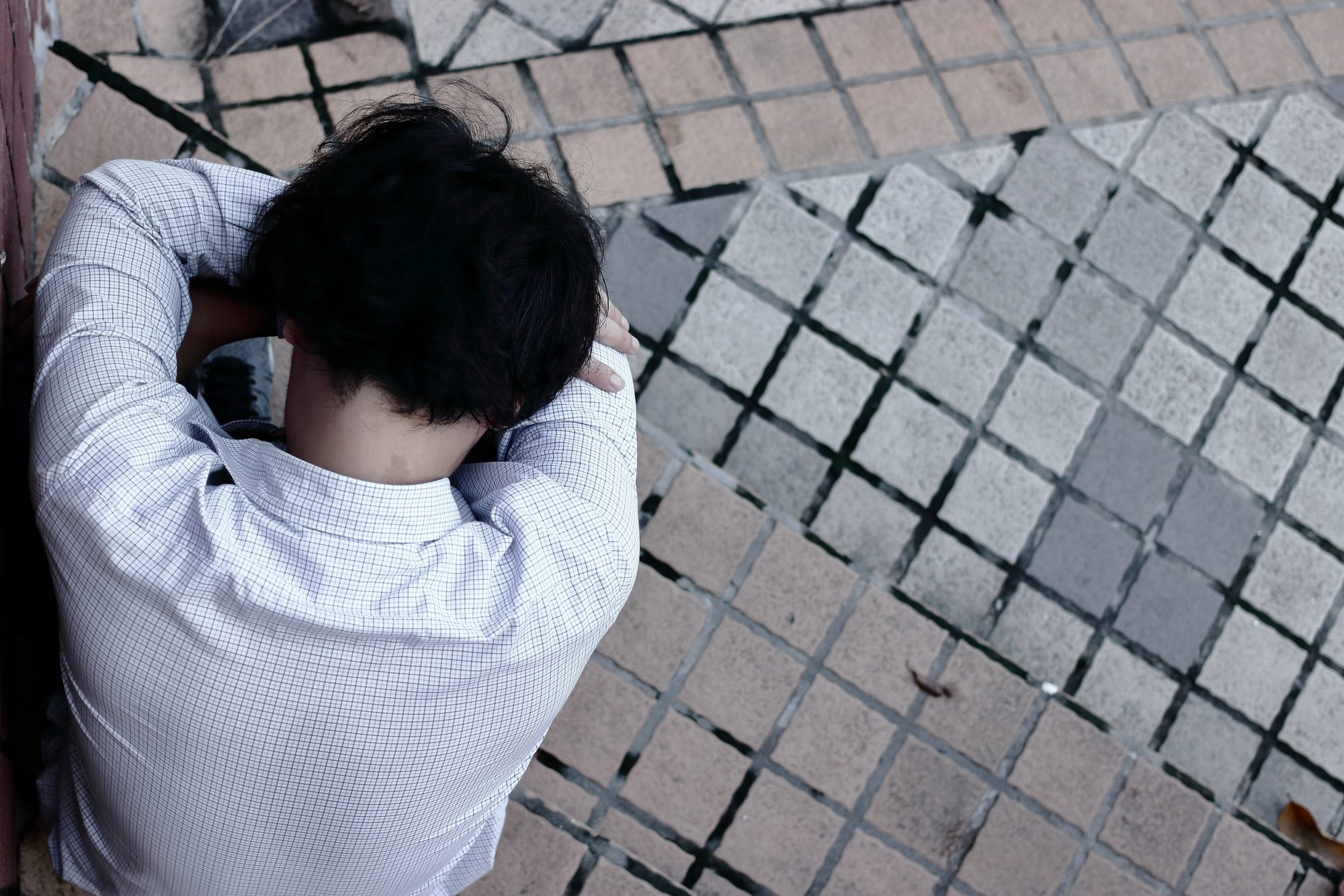 Aerial view of young Asian man in shirt with head in his hands crouched over outside due to depression and workplace violence and harm