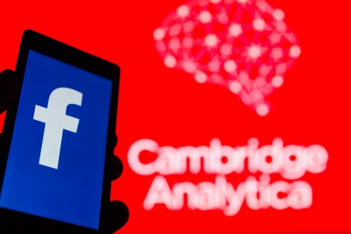 Person holding phone with Facebook app on screen in front of a red background reading 'Cambridge Analytica'