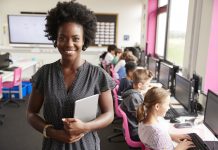 Black female teacher stood at the front of an ICT classroom. In the background, children can be seen sat working in front of laptops