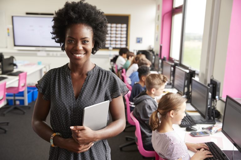 Black female teacher stood at the front of an ICT classroom. In the background, children can be seen sat working in front of laptops