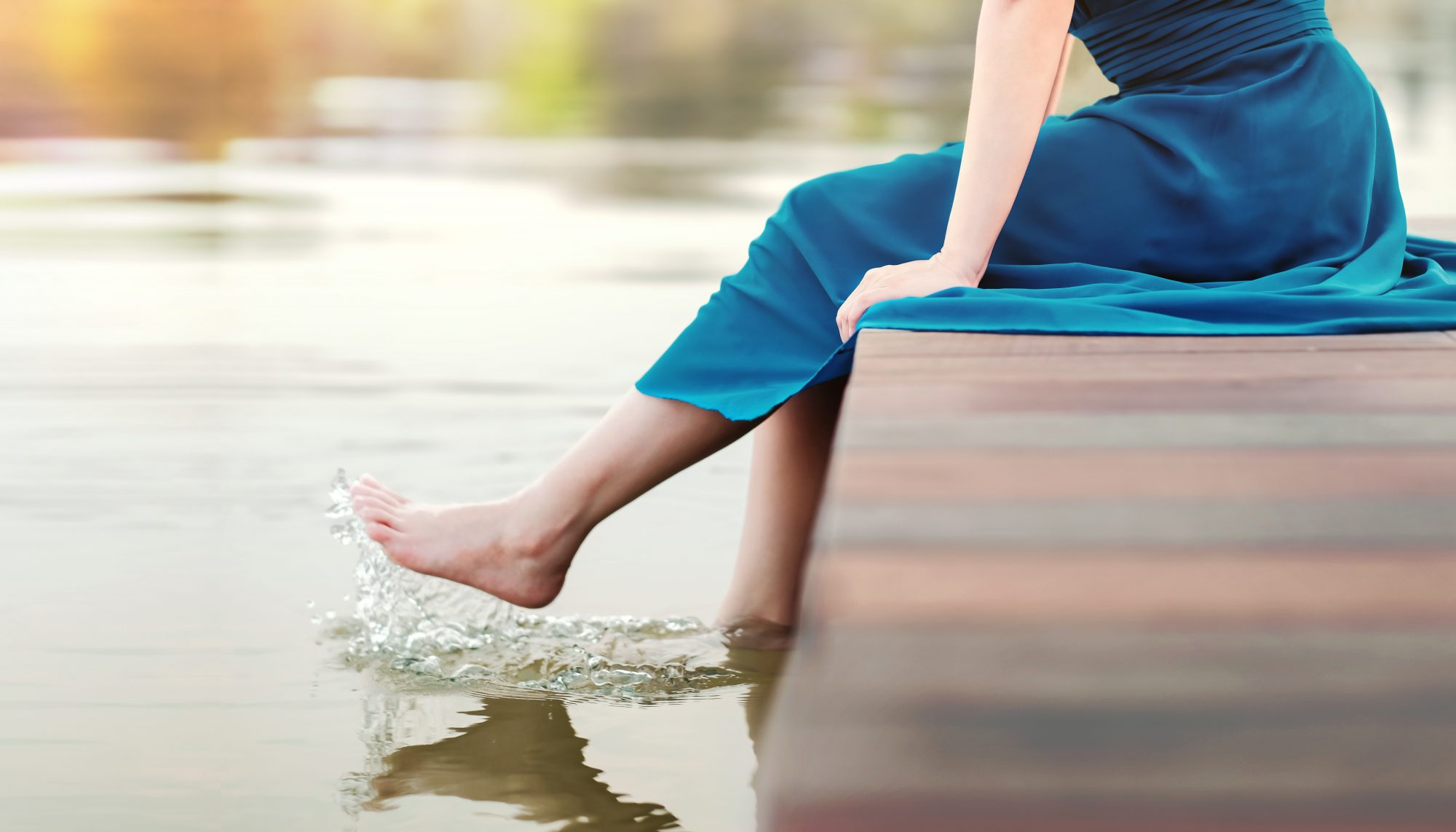 Woman in long blue dress dips her feet into lake