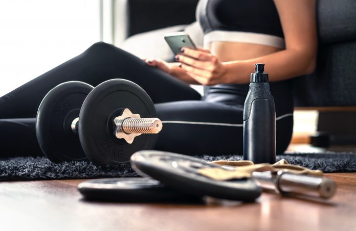 Woman in exercise gear sitting on floor behind weights using health app to guide her workout.
