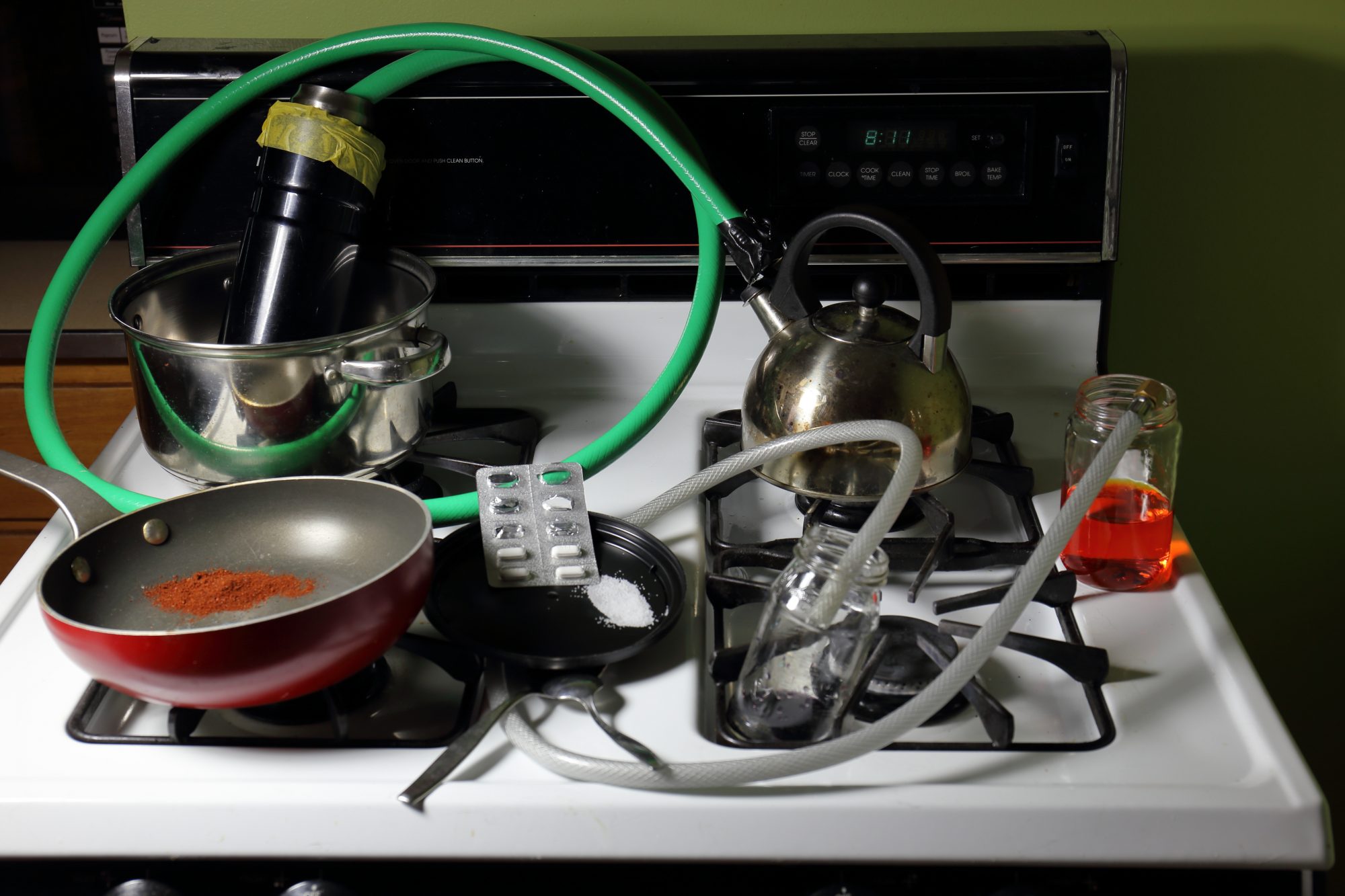 Crudely constructed meth lab on a stove top consisting of crushed nasal decongestant pills, solutions, a red powder, pots and pans, and hoses
