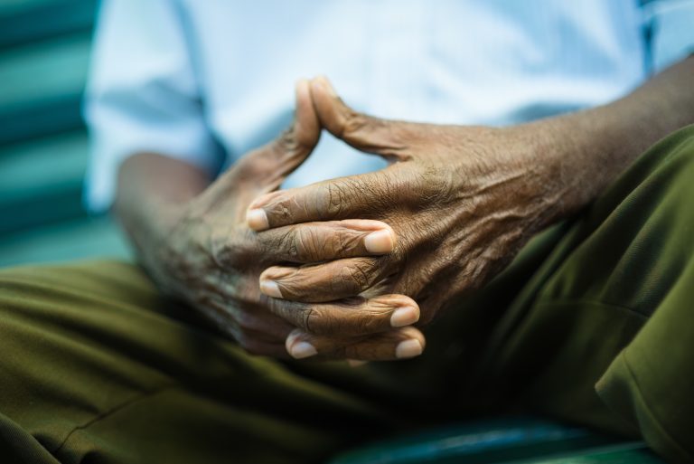 Older black man sitting on park bench with fingers intertwined, feeling pensive