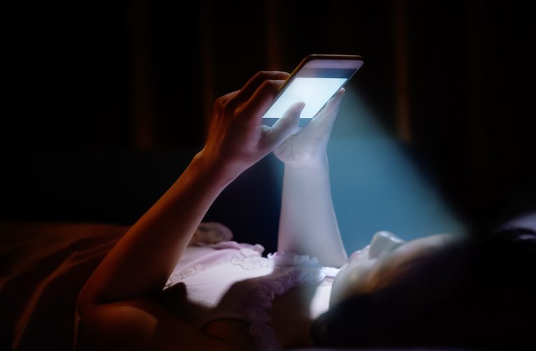 Young woman on phone in bed, blue light contributing to aging process