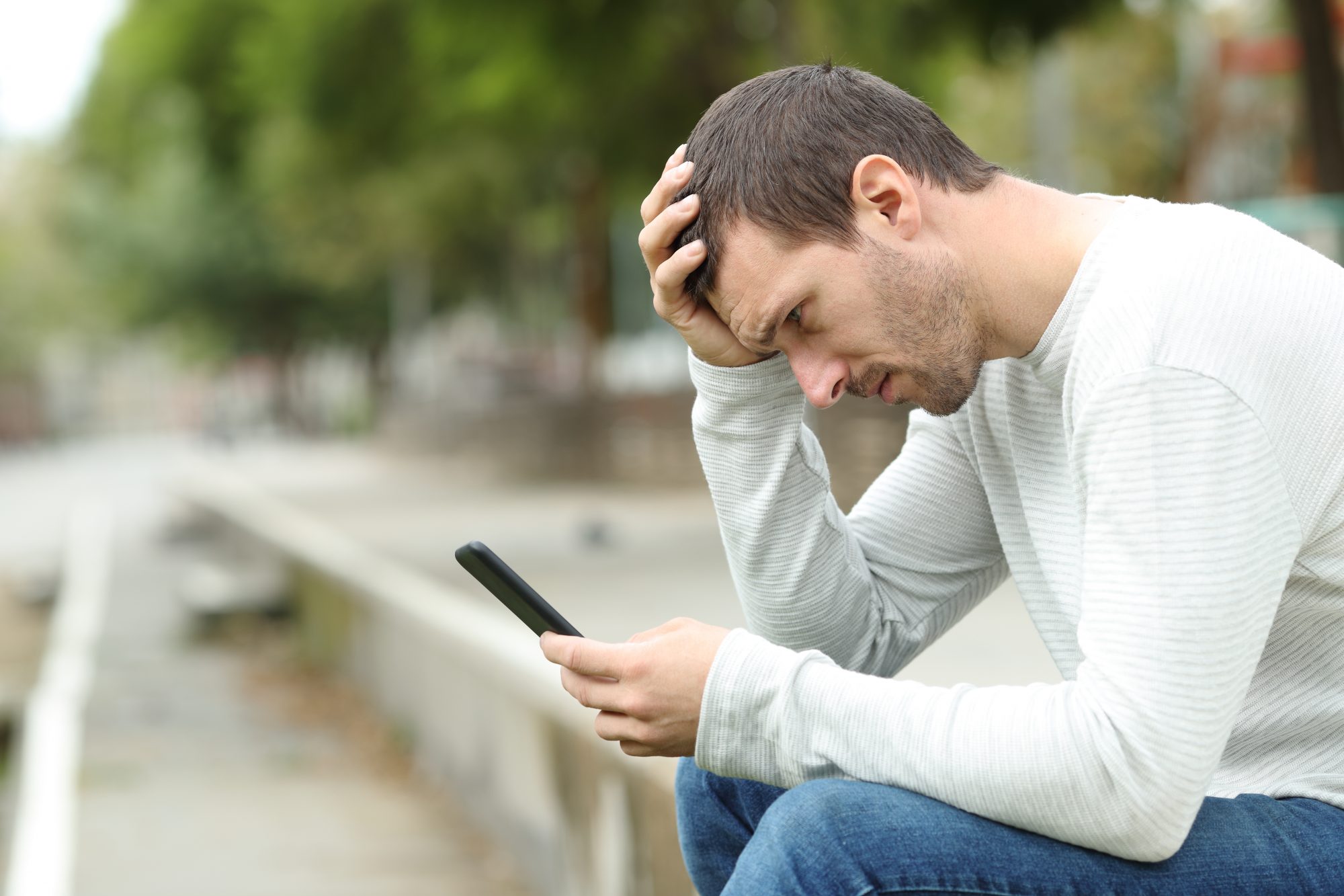 Adult white man sat on step leaning over phone as he scrolls through news.