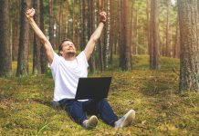 Man enjoys working remotely. sat down in forest with laptop, hands in air, expressing his enjoyment.