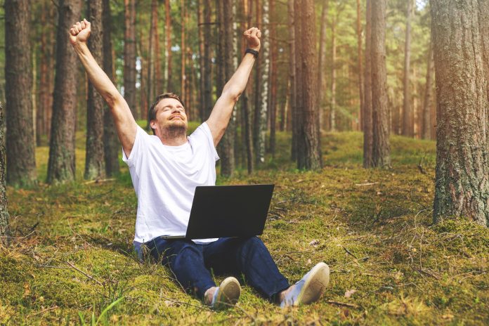 Man enjoys working remotely. sat down in forest with laptop, hands in air, expressing his enjoyment.