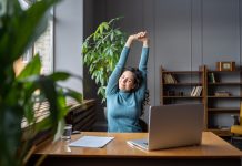 Woman stretching whilst sat at office desk, plants next to table, light coming in from window, supporting employee mental health and well-being