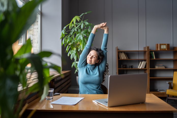 Woman stretching whilst sat at office desk, plants next to table, light coming in from window, supporting employee mental health and well-being