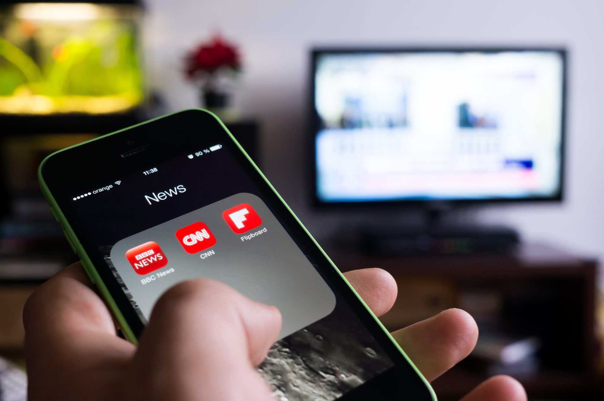 Person holding phone with news apps open. TV is visible in background