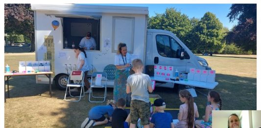 Van parked up on grassy area; adults and children sitting and standing around it participating in outreach event