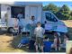 Van parked up on grassy area; adults and children sitting and standing around it participating in outreach event