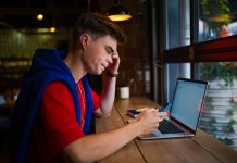 Young man working on laptop in cafe, getting distracted by phone