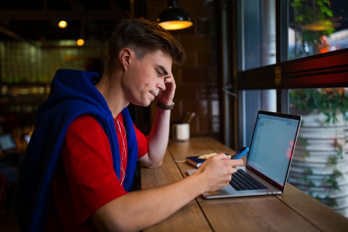 Young man working on laptop in cafe, getting distracted by phone