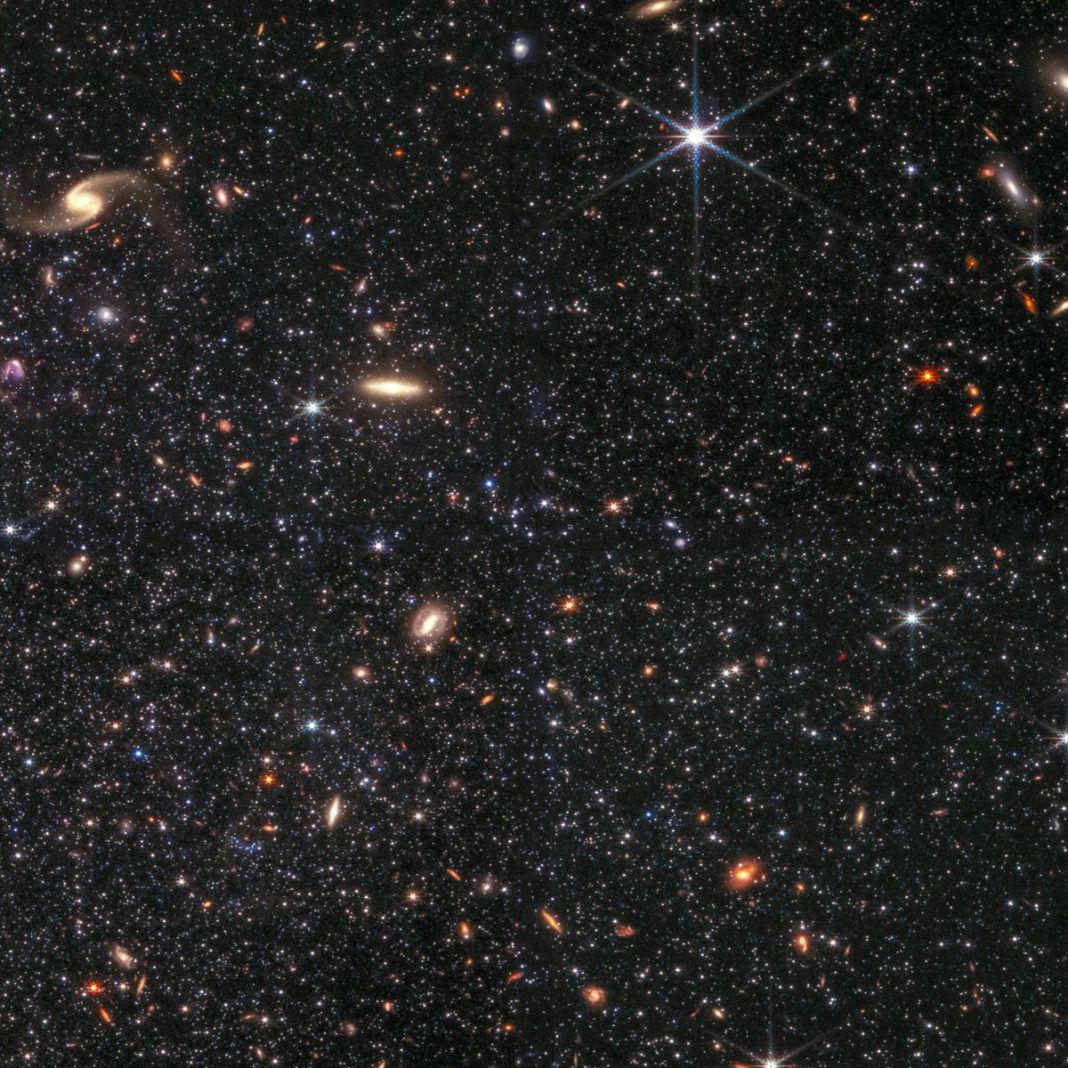 Dwarf galaxy Wolf-Lundmark-Melotte as viewed by the Webb telescope’s NIRCam instrument. Countless white stars, interspersed with yellow and orange background galaxies of various shapes, dot the black background. One prominent galaxy is a pale yellow spiral in the top left corner of the image. There is another pale yellow galaxy in the shape of a long bar closer to the center of the image. Another defining feature is a large white star with long diffraction spikes, seen just to the right of the top center.