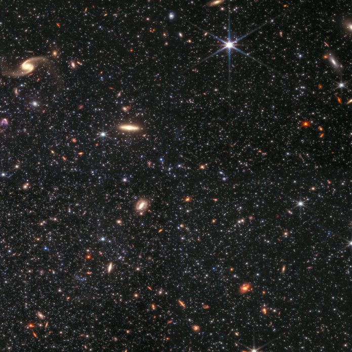 Dwarf galaxy Wolf-Lundmark-Melotte as viewed by the Webb telescope’s NIRCam instrument. Countless white stars, interspersed with yellow and orange background galaxies of various shapes, dot the black background. One prominent galaxy is a pale yellow spiral in the top left corner of the image. There is another pale yellow galaxy in the shape of a long bar closer to the center of the image. Another defining feature is a large white star with long diffraction spikes, seen just to the right of the top center.