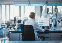 Medical Laboratory with Team of Scientists Working. Microbiologist is Using Personal Computer, Screen Shows High-Tech Concept for DNA research. High-Tech Biotechnology Research. Back View Shot