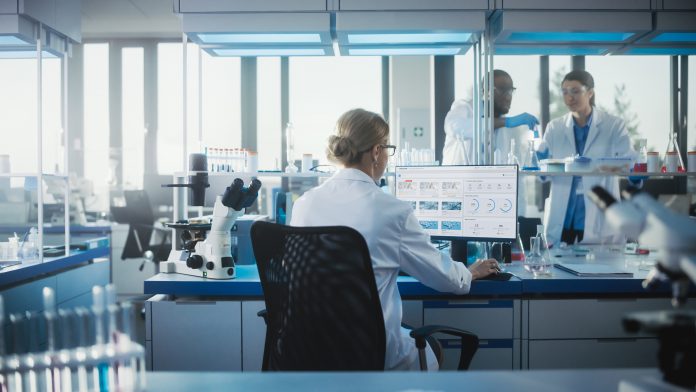 Medical Laboratory with Team of Scientists Working. Microbiologist is Using Personal Computer, Screen Shows High-Tech Concept for DNA research. High-Tech Biotechnology Research. Back View Shot