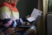 Woman In Gloves With Laptop And Bill Trying To Keep Warm By Radiator During Cost Of Living Energy Crisis