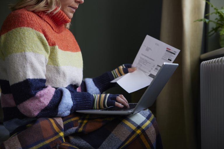 Woman In Gloves With Laptop And Bill Trying To Keep Warm By Radiator During Cost Of Living Energy Crisis