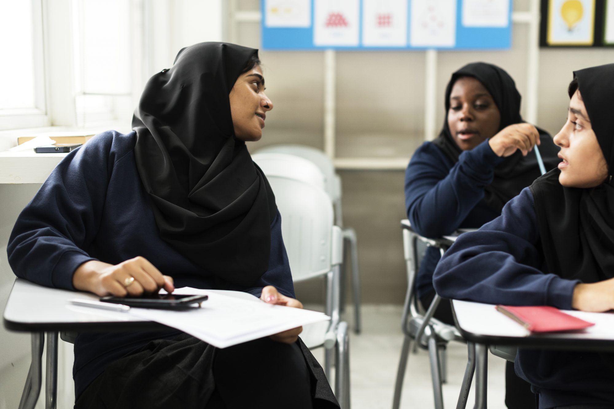 Teenage girls wearing headscarves sitting in classroom and chatting to each other 