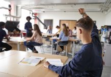 Young white male sat at desk in classroom with hand raised to attract attention of teacher