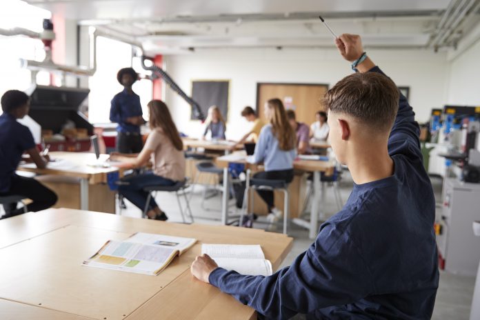 Young white male sat at desk in classroom with hand raised to attract attention of teacher