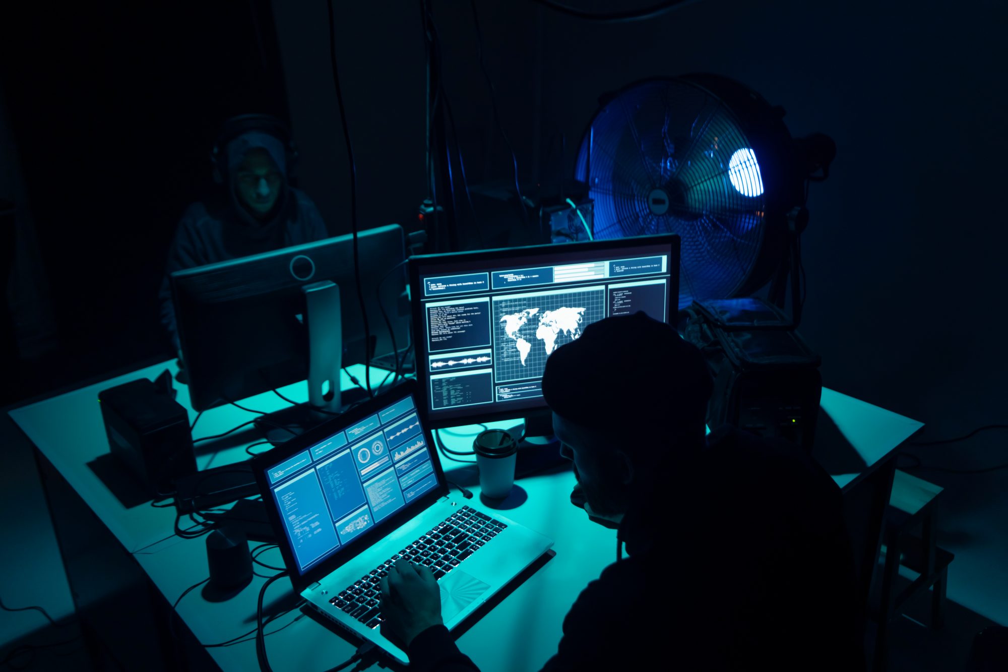 Hackers sat round table in dark room lit up by some blue lights breaking server using multiple computers and infected virus ransomware