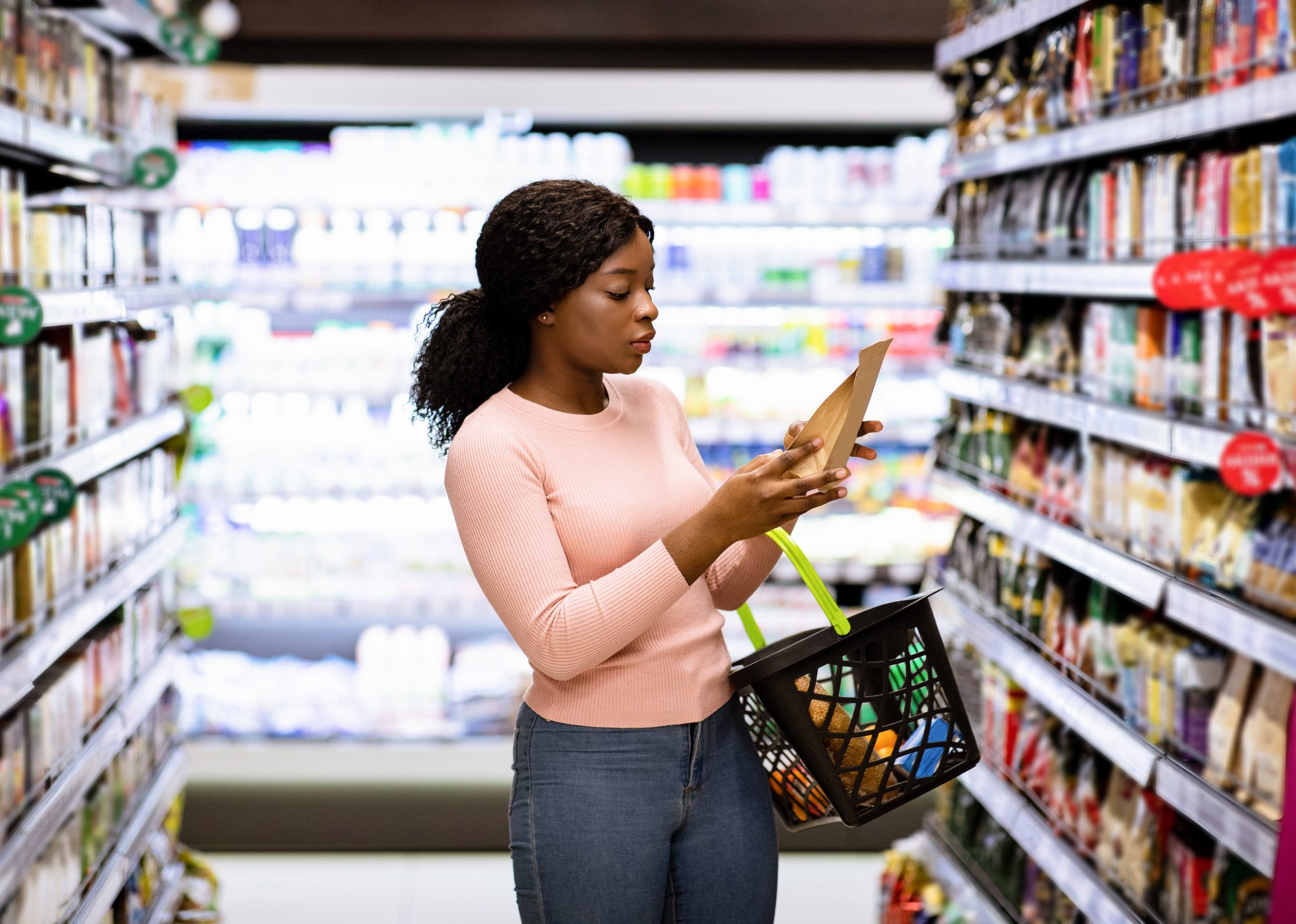 Black woman in pink jumper in supermarket aisle inspecting the packaging on back of food product