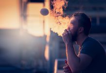 Young man with beard on blurry background sunset sky, making soap bubbles smoke inside with the aid of vape