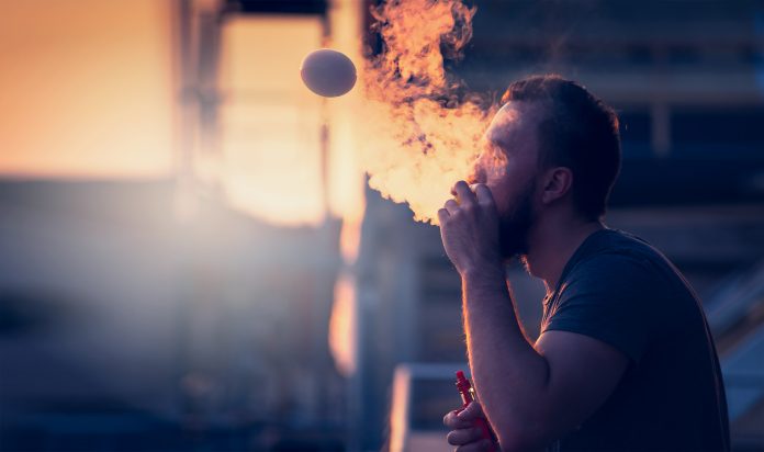 Young man with beard on blurry background sunset sky, making soap bubbles smoke inside with the aid of vape