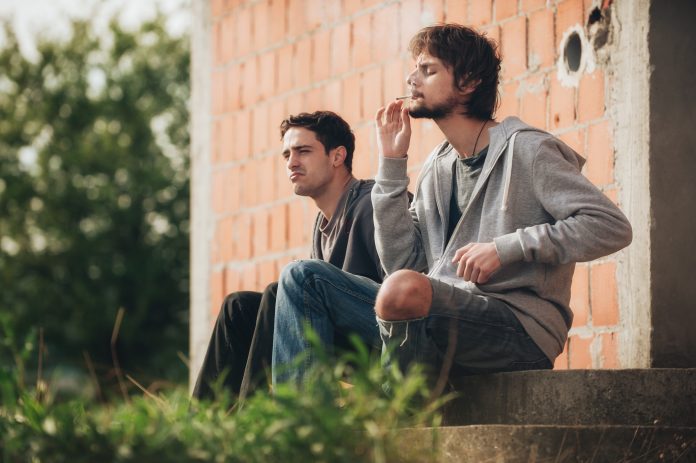 Two young men smoking cannabis, sat outside amongst greenery