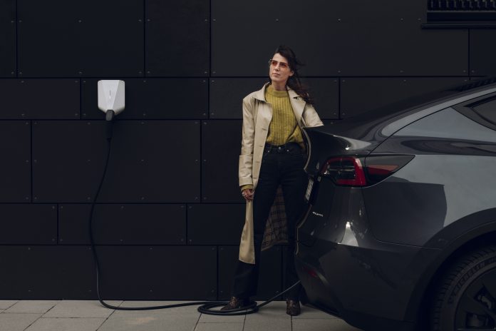 Woman in long coat stood against black wall charging up her electric vehicle
