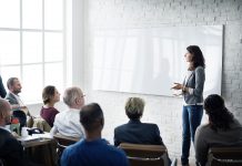 Business leader giving talk to conference room, woman standing up in front of whiteboard, everyone one else sat down in front of her