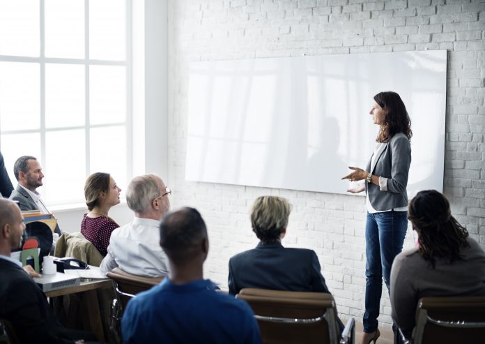 Business leader giving talk to conference room, woman standing up in front of whiteboard, everyone one else sat down in front of her