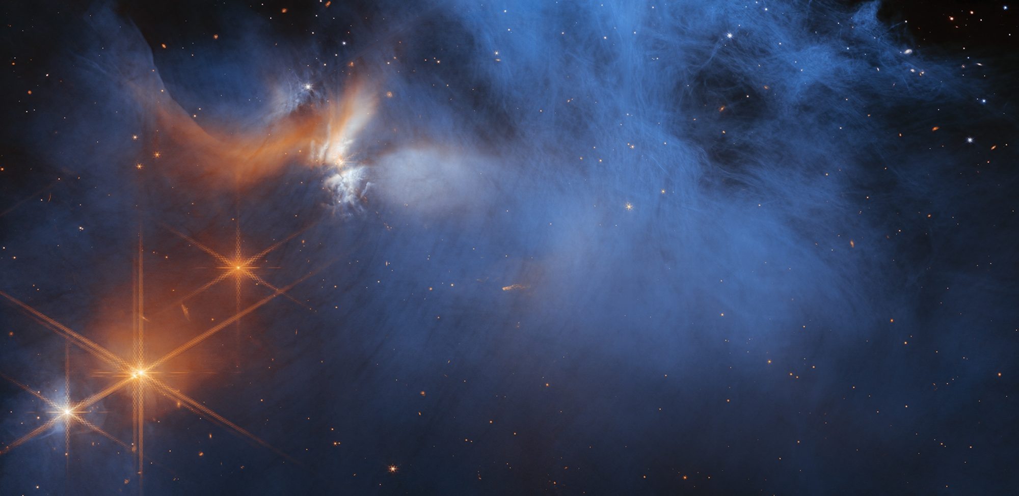 This image by NASA’s James Webb Space Telescope’s Near-Infrared Camera (NIRCam) features the central region of the Chamaeleon I dark molecular cloud, which resides 630 light years away. The cold, wispy cloud material (blue, center) is illuminated in the infrared by the glow of the young, outflowing protostar Ced 110 IRS 4 (orange, upper left). The light from numerous background stars, seen as orange dots behind the cloud, can be used to detect ices in the cloud, which absorb the starlight passing through them
