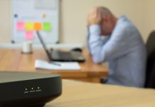 Close up of domestic broadband router showing lack of signal orange no internet light. In background home working man on computer with head in hands