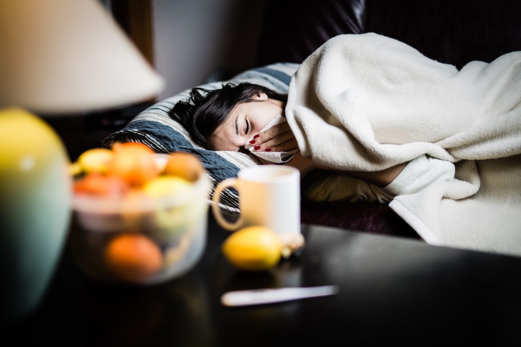 Sick woman in bed,calling in sick,day off from work.Thermometer to check temperature for fever.Vitamins and hot tea in front.Flu.Woman Caught Cold.Virus.Sick woman laying in bed under wool blanket