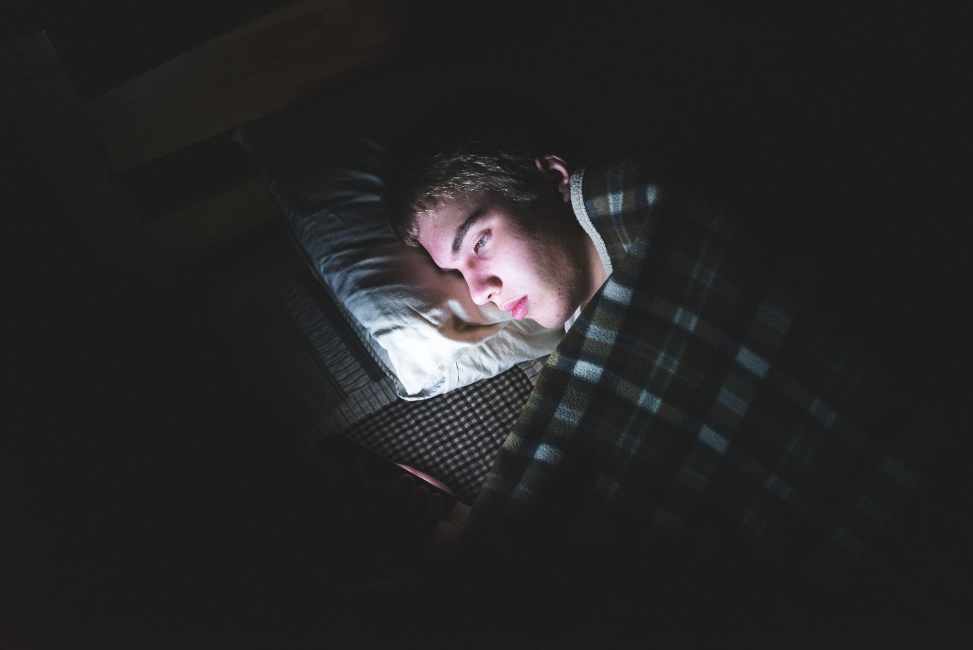 The image displays a depressed teenager browsing the internet on his mobile phone. He is lying on his bed in the dark.