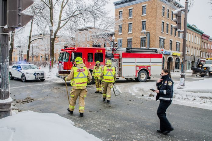 Firemen using broom to clean street corner from debris after car accident during winter day in Quebec city. Fire truck behind and police officer passing by