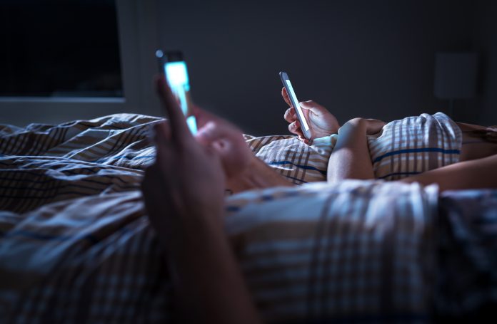 Bored distant couple ignoring each other lying in bed at night while using mobile phones. Smartphone addict. Obsessed and distracted man and woman texting. Addiction to social media and technology.