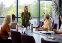 A wide-angle view of a group of a woman getting together for a business meeting in their place of work in Hexham. The woman in the green blouse is leading the meeting and is encouraging the others to take part.