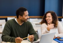 The couple smiles and talks together before their evening class. He has his laptop open and ready and she has her smart phone on the desk in front of her.