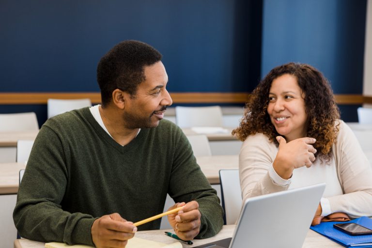 The couple smiles and talks together before their evening class. He has his laptop open and ready and she has her smart phone on the desk in front of her.